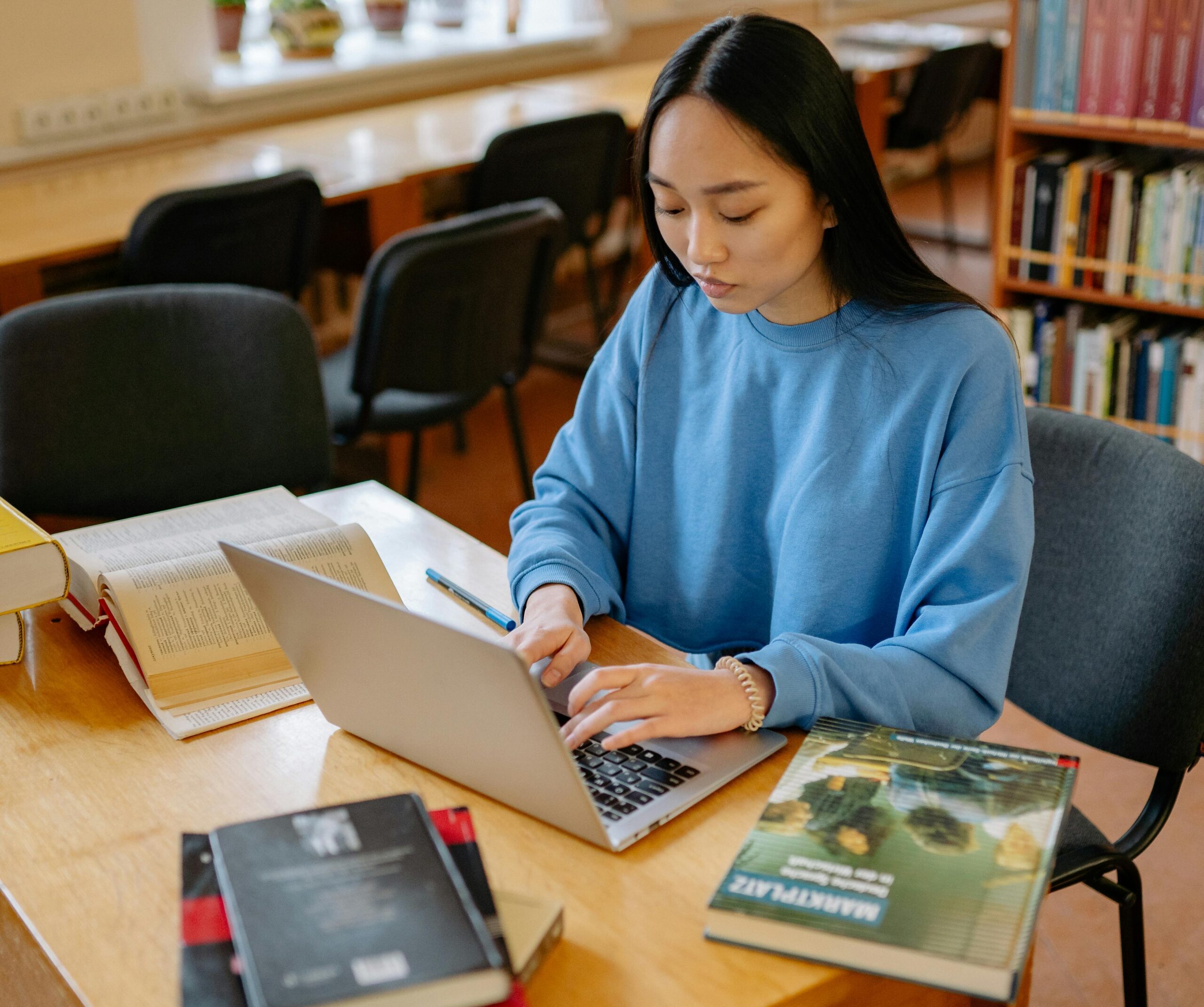 Asian woman typing on a laptop surrounded by books in a library setting.